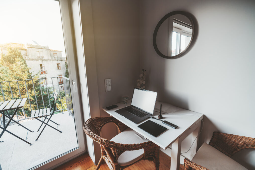 View of a cozy nook in a residential apartment with a work-table near the balcony with a beautiful view; on the table there are: opened laptop with a blank screen, multimedia adaptor, graphic tablet