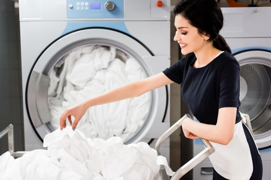 cheerful maid looking at bed sheets while standing near washing machines