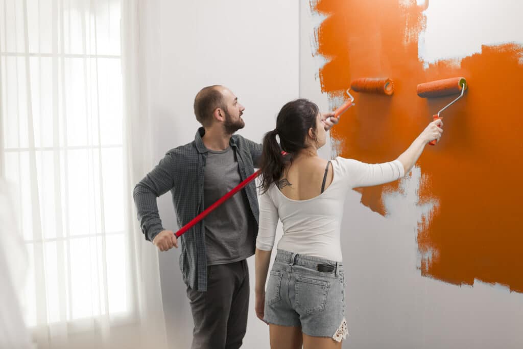Confident couple using orange color paint on walls to redecorate apartment room during interior design renovation. Doing housework redecoration with painting tools, ladder and DIY equipment.