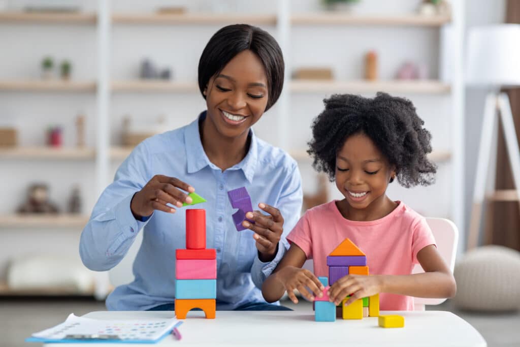 Friendly female teacher pretty young african american woman playing with curly little girl at daycare, making various constructions from colorful wood blocks, having conversation and smiling
