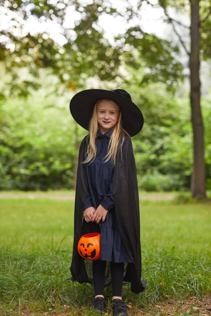 Vertical full length portrait of smiling teenage girl dressed as witch posing outdoors and holding Halloween bucket