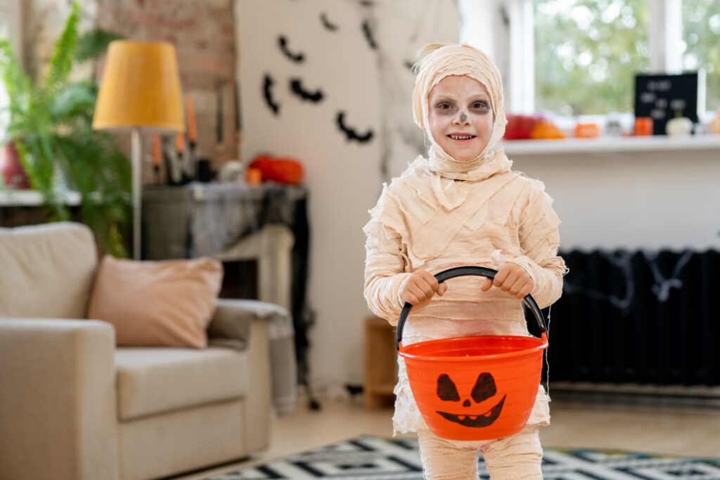 Portrait of smiling little mummy boy wrapped up into bandages holding candy bucket for trick-or-treating in living room with Halloween decorations
