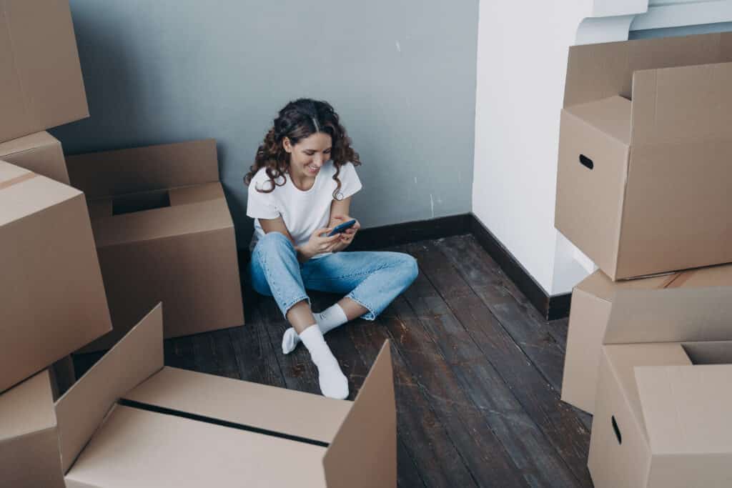Smiling female holding smartphone, choosing moving company for relocation, sitting on the floor with carton boxes in vacated apartment. Girl selects a reputable mover for removal things.